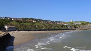 A coastal beach with a promenade, houses on a hillside, and a lighthouse in the background.