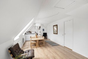 A modern loft apartment with a dining area, a black leather chair, a wooden dining table, a potted plant, and a kitchen in the background.