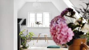 Ein Zimmer mit weißer Decke, einem Fernseher links, einem Fenster mit weißen Jalousien, einem kleinen Tisch und einer Topfpflanze.
