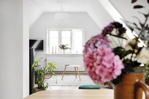 Ein Zimmer mit weißer Decke, einem Fernseher links, einem Fenster mit weißen Jalousien, einem kleinen Tisch und einer Topfpflanze.