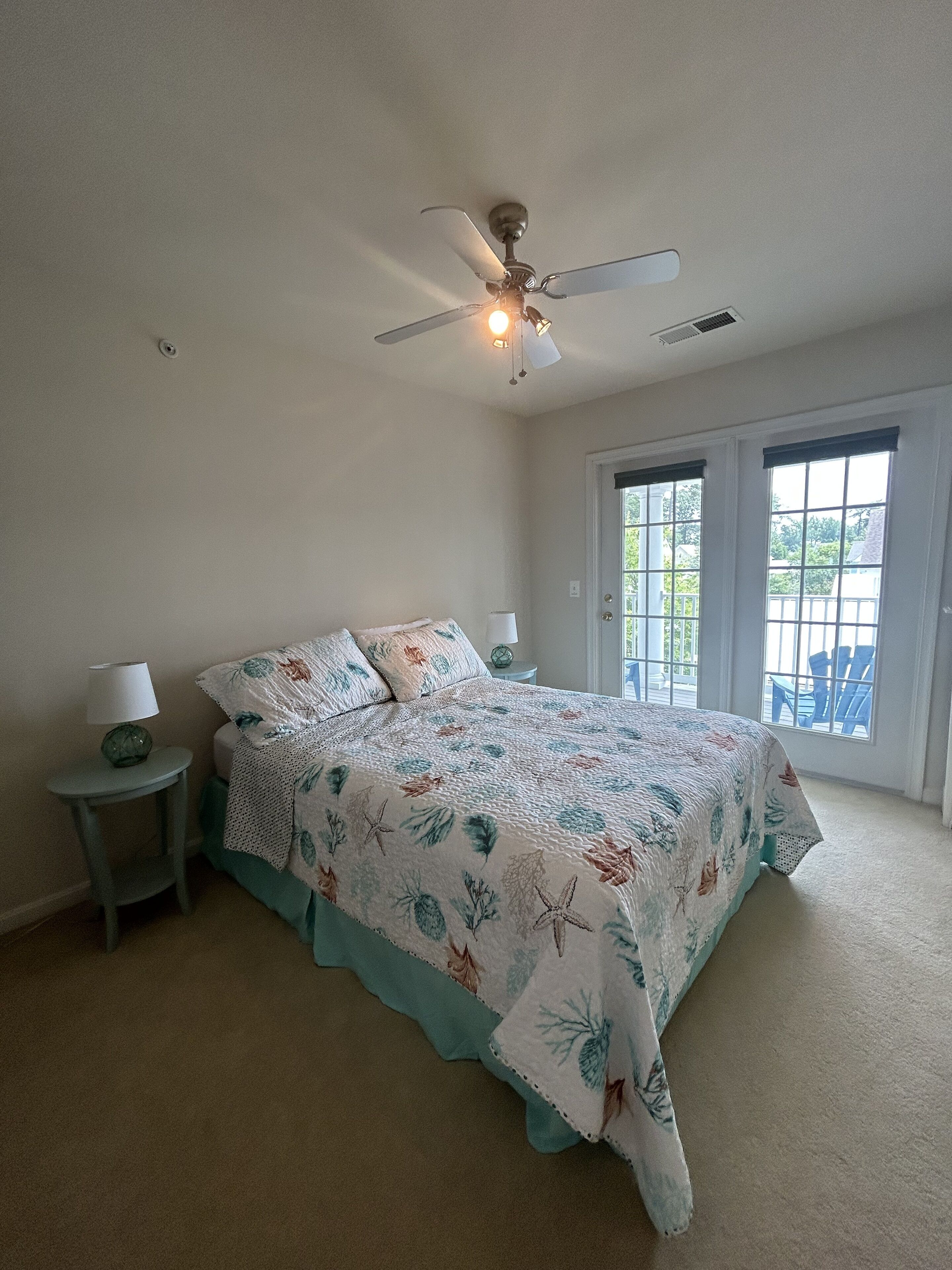 A bedroom with a bed, a ceiling fan, a nightstand with a lamp, and a door leading to an outdoor area.