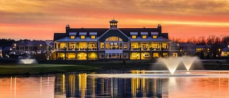 A large, illuminated building with a central entrance and multiple windows, situated across a body of water with fountains.