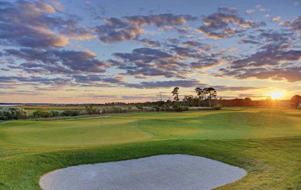 A golf course with a sand bunker, trees, and a sunset in the background.