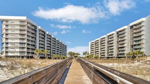 A wooden boardwalk leading to a modern hotel with multiple balconies and a clear blue sky.