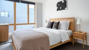 A modern bedroom with a large bed, a wooden nightstand, a bedside lamp, and a window with blinds. - Stunning 2BR in East London, nr Old Street stasion, by Blueground (London)