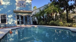 En swimmingpool omgivet af et hus med veranda og palmer.