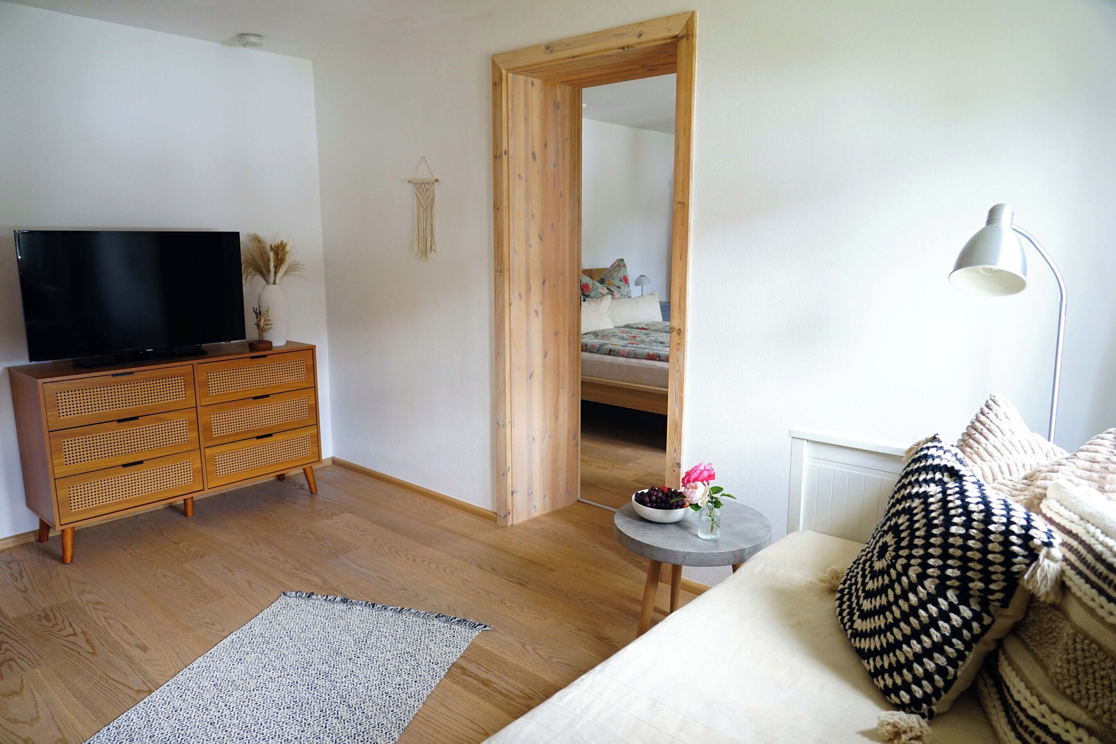 Ett rum med en byrå i trä, en tv, ett litet runt bord med en vas och blommor, en säng med ett mönstrat kuddöverdrag och en spegel som reflekterar sovrummet.