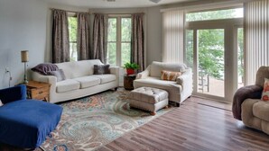 Een woonkamer met een witte bank, een blauwe fauteuil en een beige pouf. Op de vloer ligt een tapijt met een patroon en er staat een kleine tafel met een lamp.