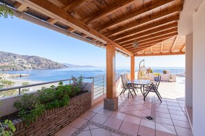 Una terraza techada con vista al mar, una mesa con sillas y una jardinera de ladrillo con plantas.