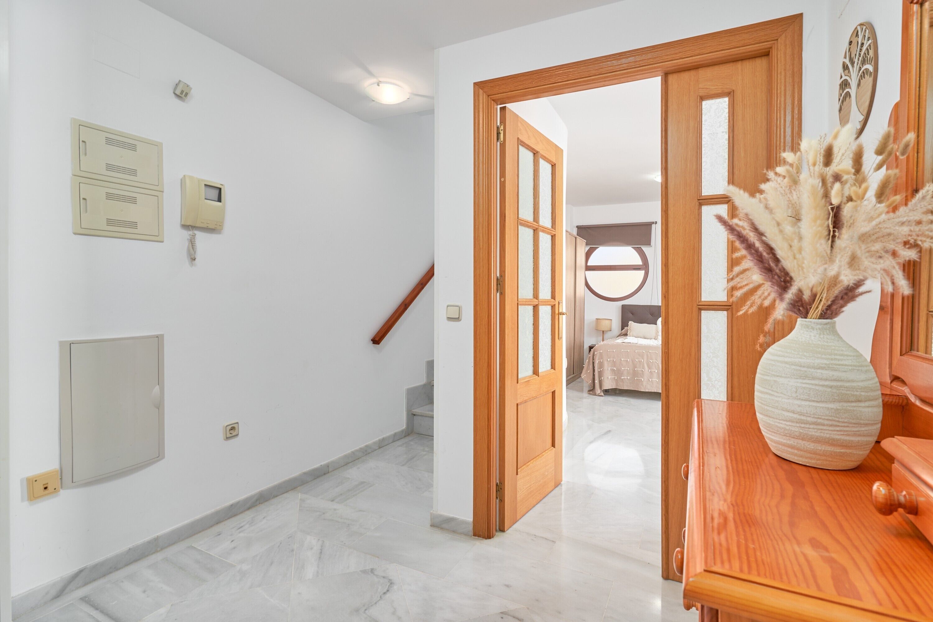 Un pasillo con una puerta de madera que da a un dormitorio, un jarrón con plumas de miscanthus y un piso de mármol.