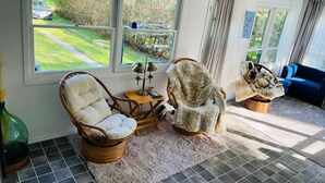 A cozy living room with wicker chairs, a fluffy rug, and a view of a green garden through large windows. - Lake Huron Getaway: Cozy Cottage, Private Access, Second-Row Location (Dashwood)