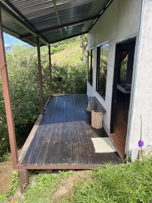 A covered wooden deck with a bench, a small table, and a window leading to a bedroom. - Cabañas Colibrí Tayutic (Tayutic)