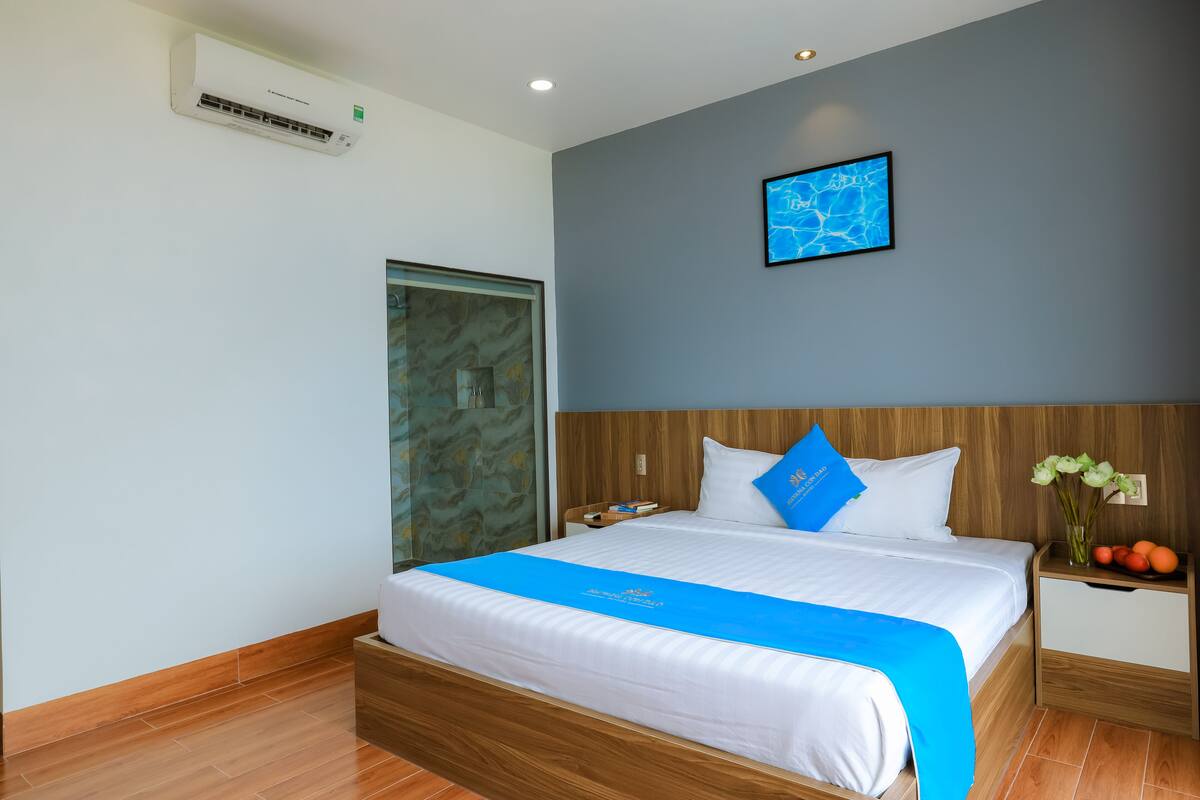 Quarto de hotel moderno com cama de madeira, tapete decorativo azul, mesa de cabeceira com um vaso de flores e ar condicionado fixado na parede.