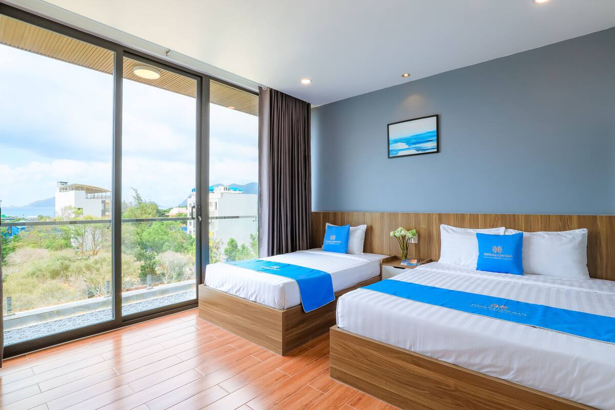 Quarto de hotel com duas camas, uma janela grande e vista da cidade, e uma decoração em azul e branco.