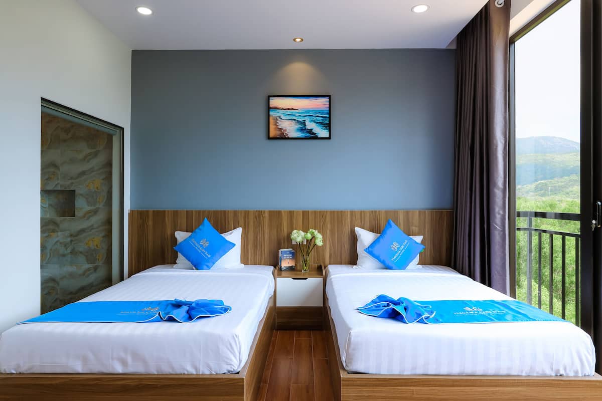 Quarto de hotel com duas camas, um quadro de uma paisagem, uma mesa de cabeceira com um vaso de flores e vista para o verde através de uma janela.
