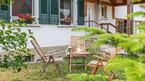 Een terras met houten stoelen en een tafel, een raam met groene luiken en een bloeiende plant in een pot.