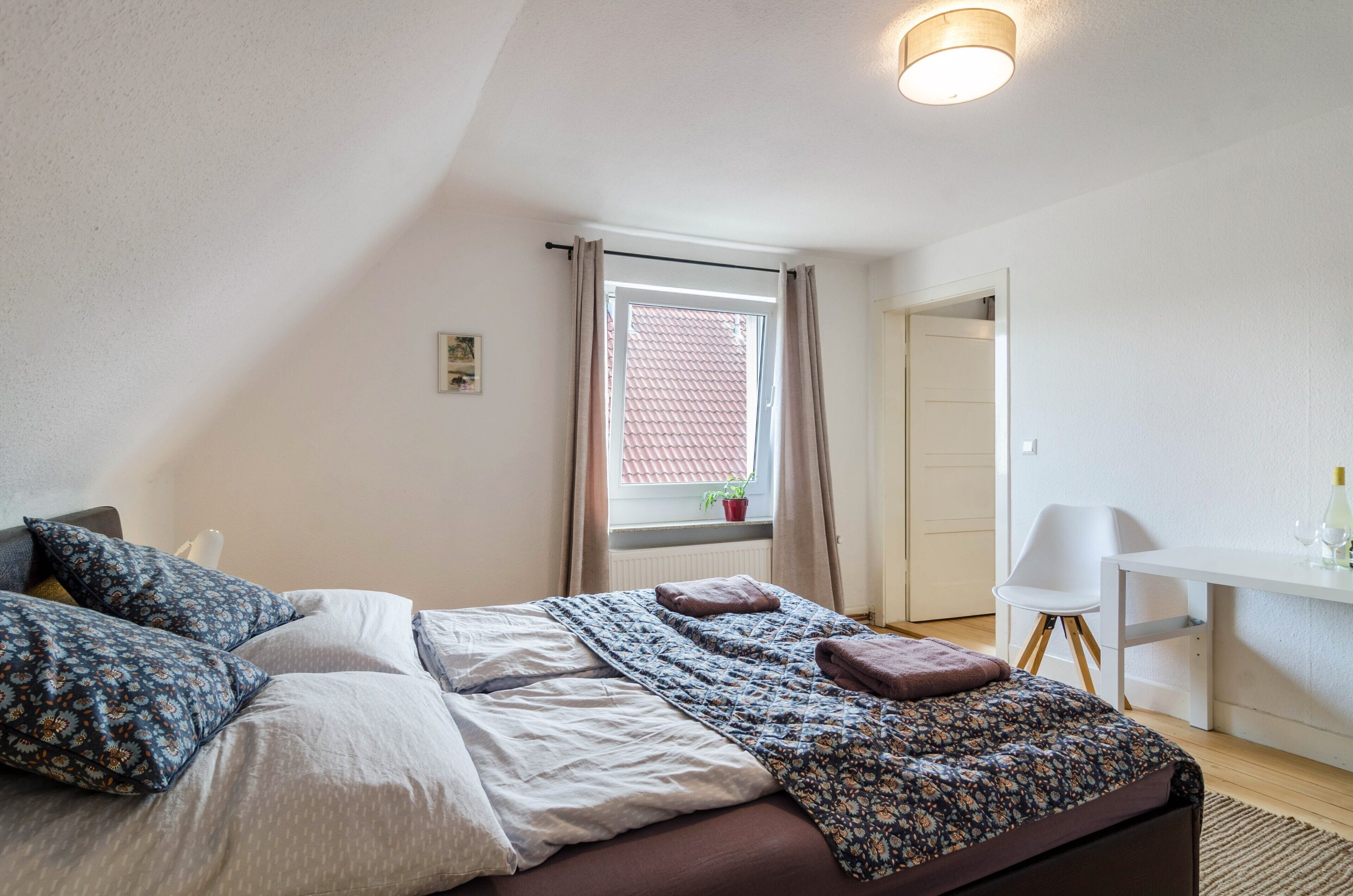 Een slaapkamer met een bed, een bureau, een stoel, een raam met gordijnen en een plant.