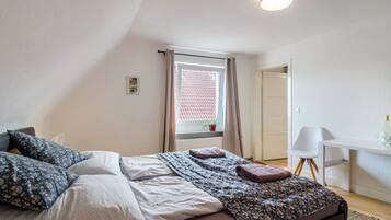 Een slaapkamer met een bed, een bureau, een stoel, een raam met gordijnen en een plant.