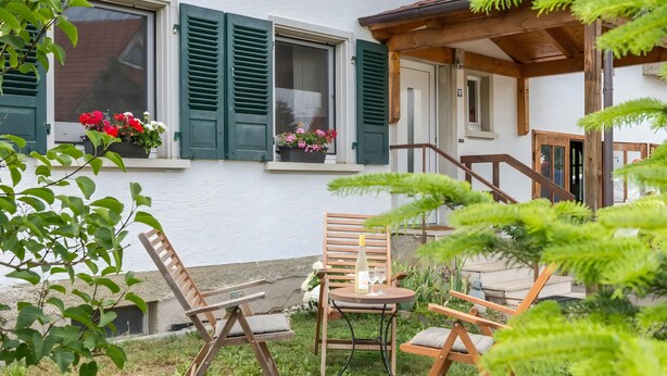 Un patio con sillas y mesa de madera, una ventana con contraventanas verdes y una planta en maceta con flores.