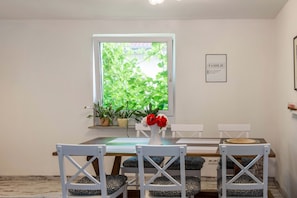 Un espace repas comprenant une table, des chaises, un vase avec des fleurs, des plantes en pot et un panneau encadré accroché au mur.