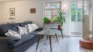 Un salón con un sofá gris, una mesa de centro de vidrio, una planta en maceta y una puerta que da al exterior.