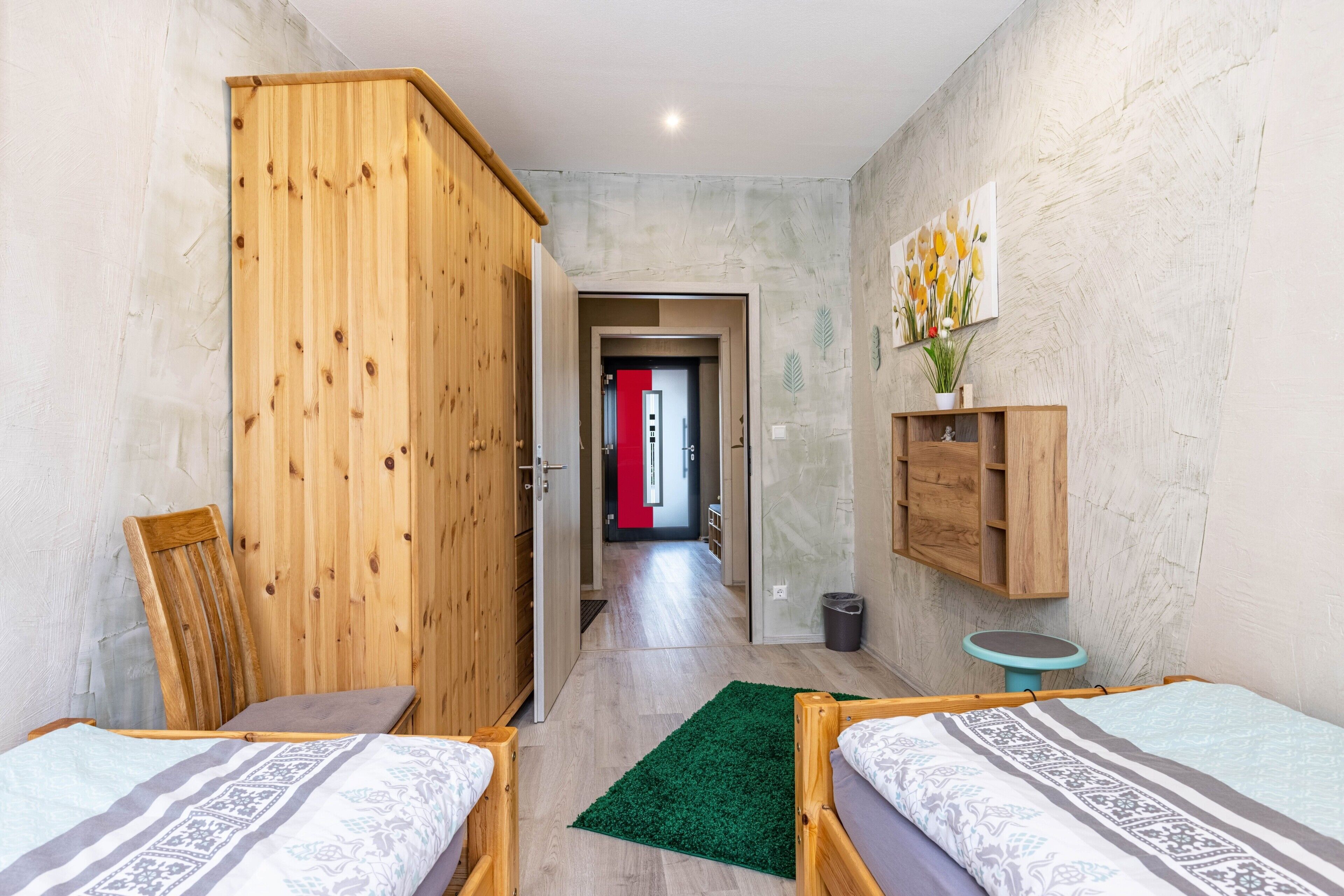 A room with two beds, a wooden wardrobe, a green rug, a small round table, and a wall-mounted shelf with a plant.