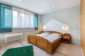 Ein Schlafzimmer mit einem hölzernen Bett, einem Nachttisch, einem grünen Teppich, einem Fenster mit durchsichtigen Vorhängen und einer Wandleuchte.