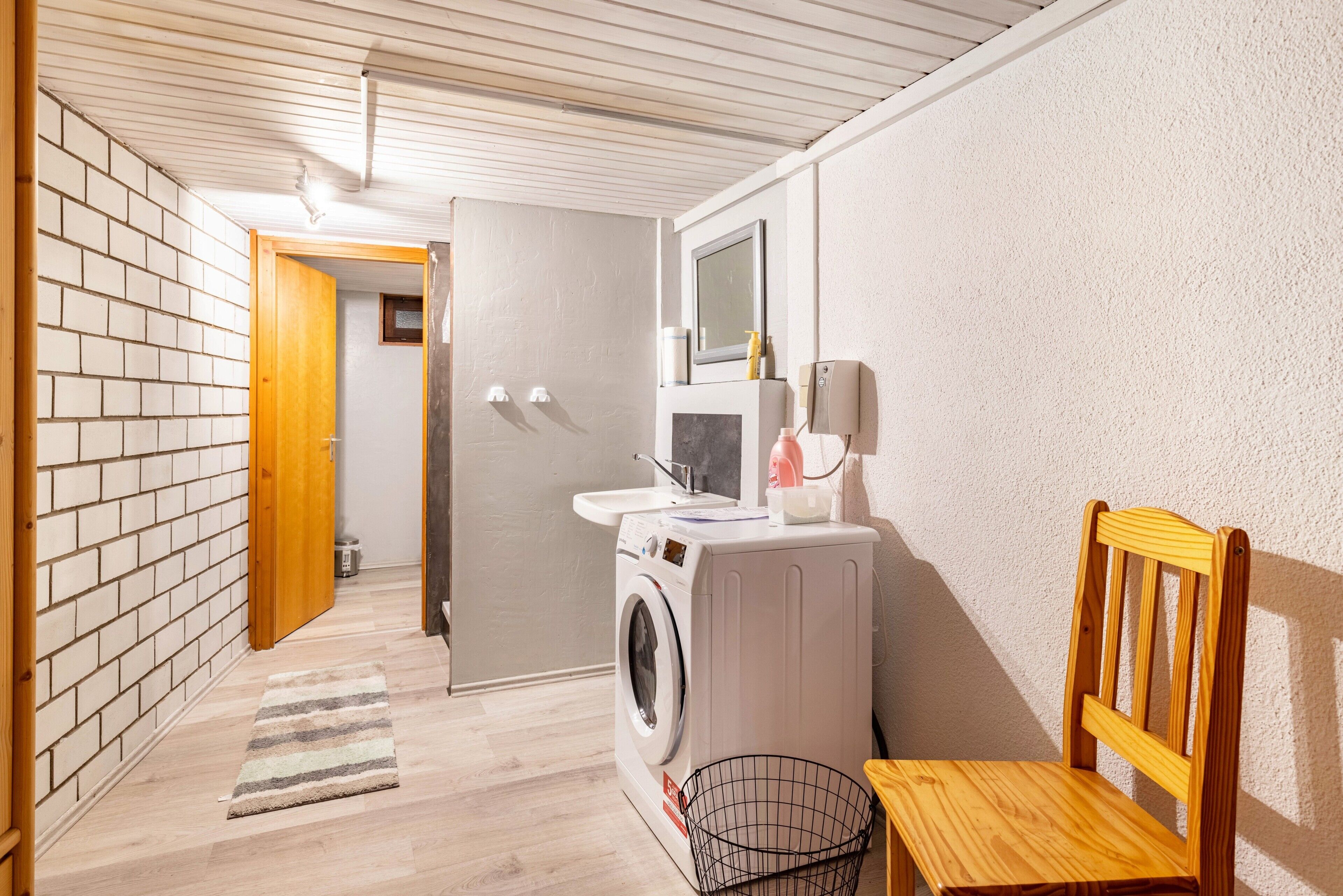 A laundry room with a washing machine, a sink, a mirror, and a wooden chair.