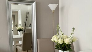 Una habitación con un espejo grande, una mesa blanca con diseño floral, un florero con flores y una lámpara de pie.