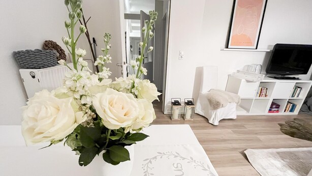Una sala de estar con un sofá blanco, un televisor, una estantería y un arreglo floral sobre una mesa.