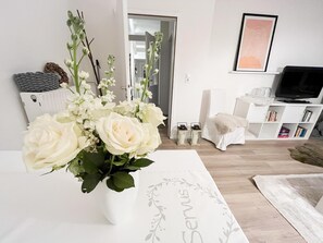 Un salon avec un canapé blanc, un téléviseur, une bibliothèque et un arrangement floral sur une table.