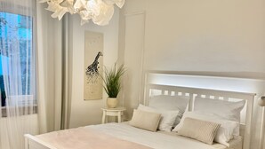 Una cama blanca con cabecera, una mesita de noche con una planta y una obra de arte colgada en la pared.