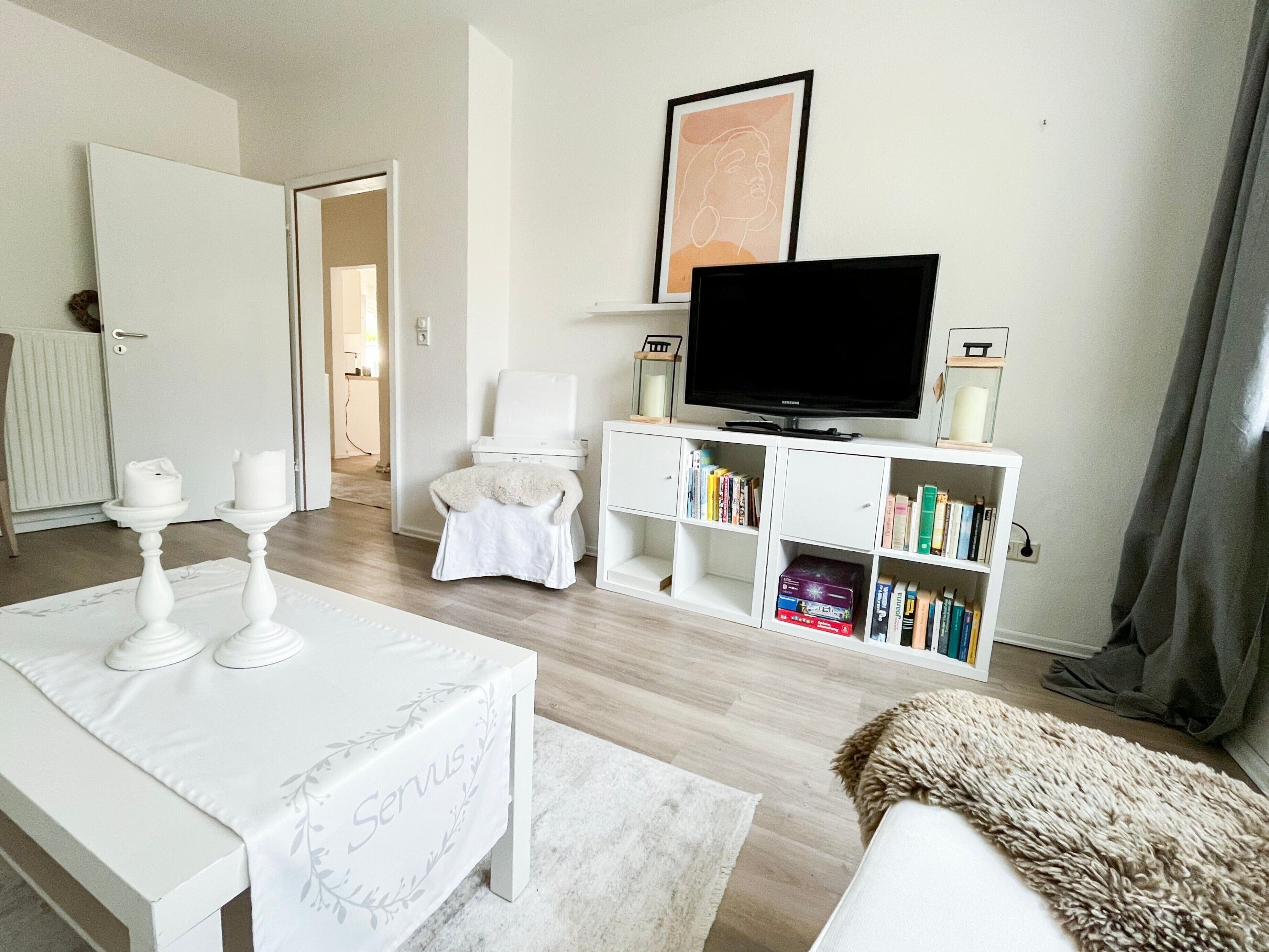 A modern living room with a white sofa, a white table with candles, a television, and a bookshelf.