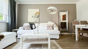 Una sala de estar moderna con un sofá blanco, una mesa de centro, una lámpara de pie y una zona de comedor con una mesa y sillas.