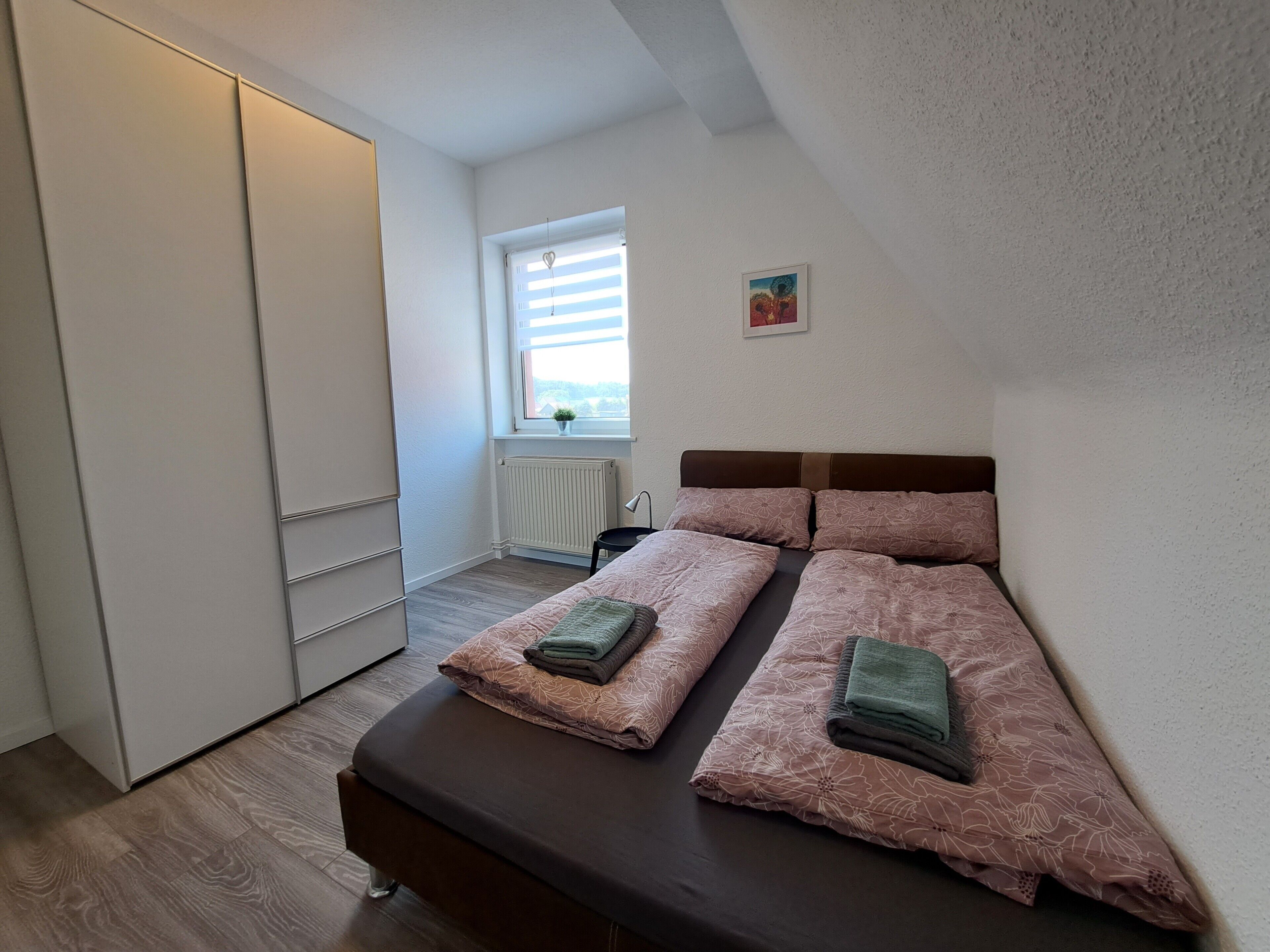 Une chambre à coucher avec un lit, un radiateur, une fenêtre avec des stores et une petite plante posée sur le rebord.
