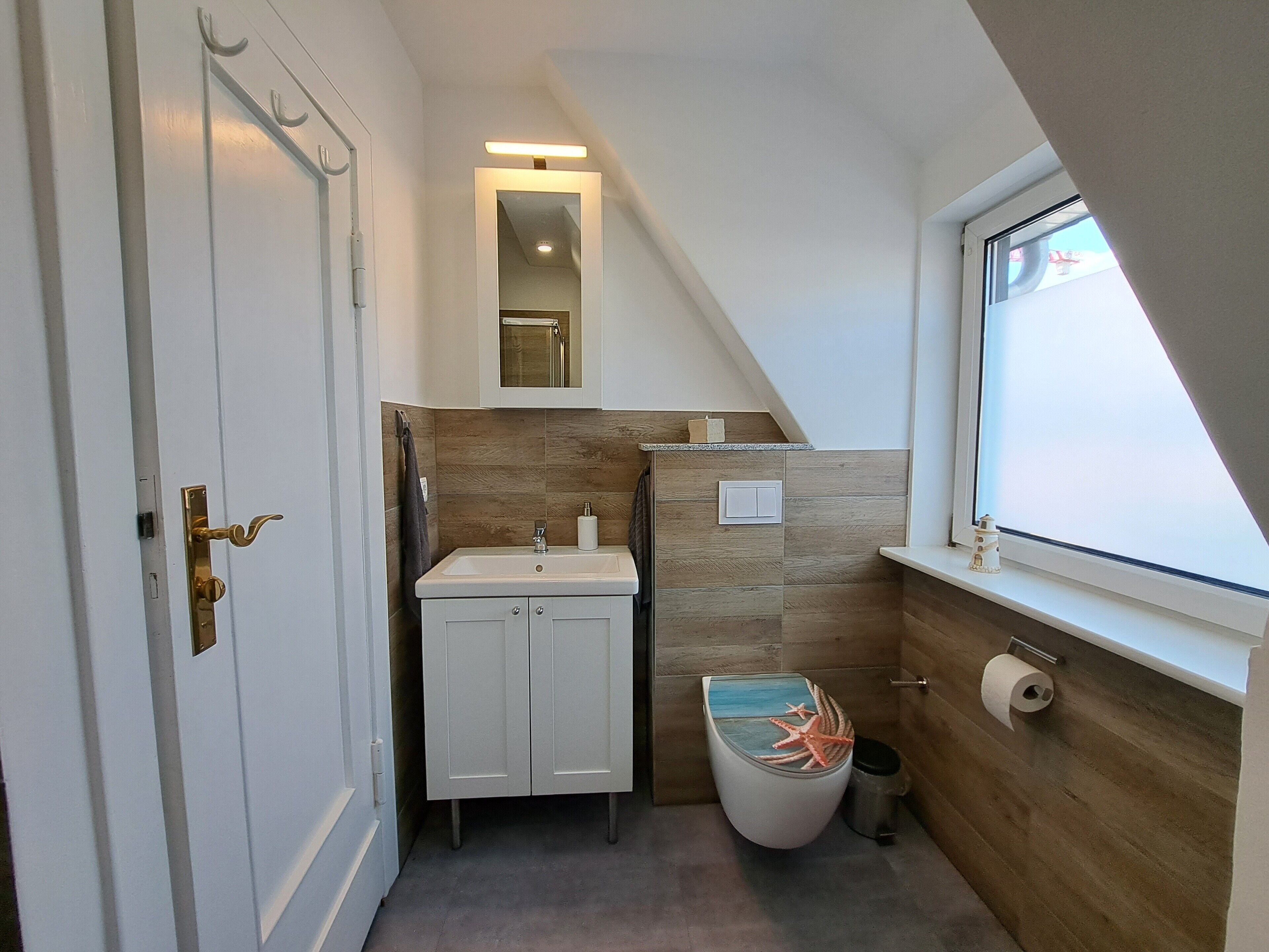 Een badkamer met een witte wastafel, een toilet met een decoratieve afdekking, een spiegel boven de wastafel en een raam met uitzicht.
