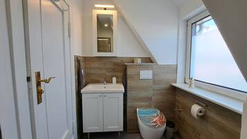 Een badkamer met een witte wastafel, een toilet met een decoratieve afdekking, een spiegel boven de wastafel en een raam met uitzicht.