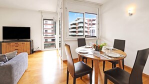 A modern living room with a flat-screen TV, a grey sofa, a wooden dining table set for four, and a large window with a view of a building.