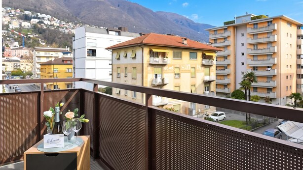 A balcony with a table, a bottle, and glasses, overlooking a cityscape with buildings and mountains.