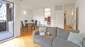 A modern living room with a grey sofa, a dining table with chairs, a kitchen in the background, and a balcony with a view of buildings.