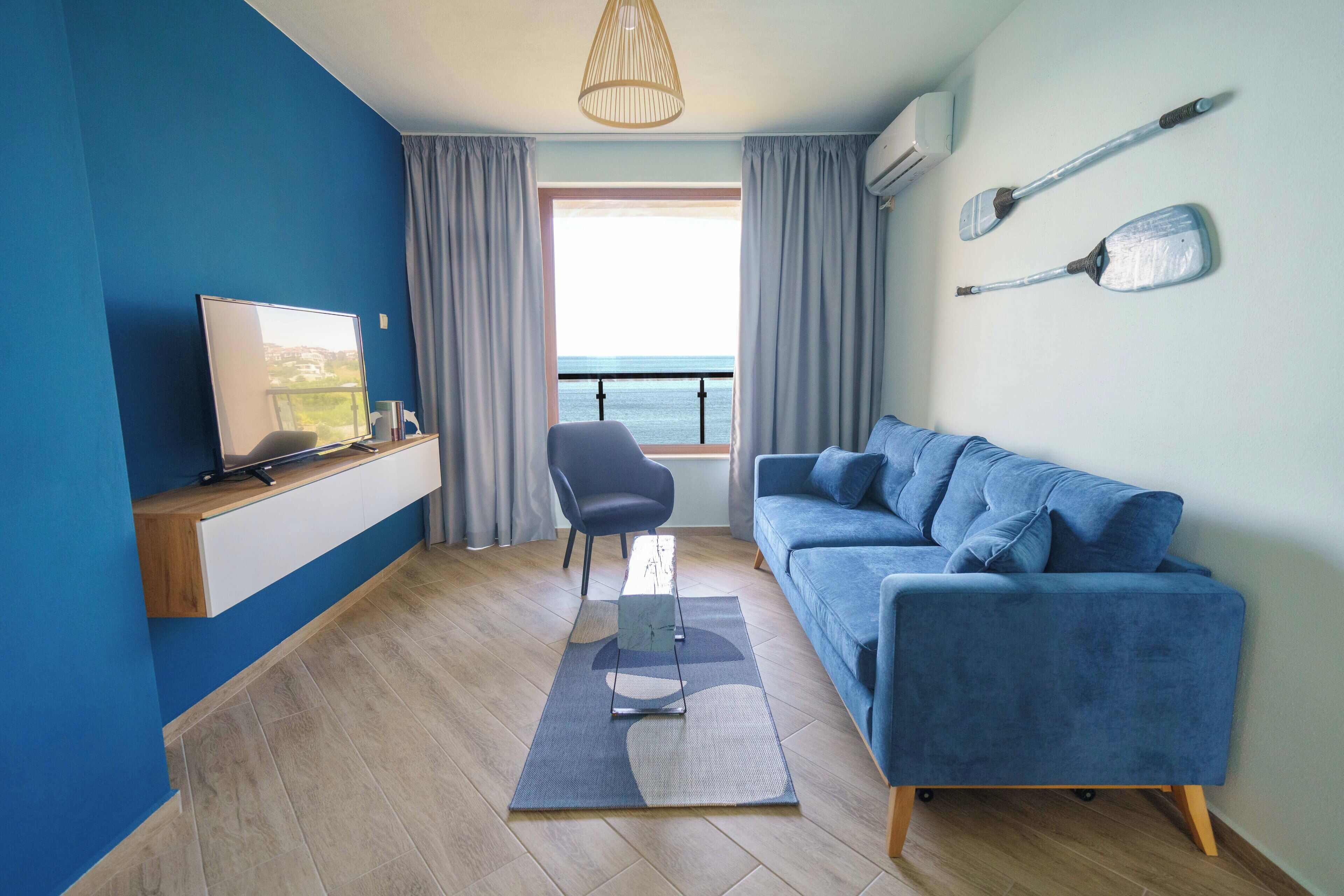 A modern living room with a blue sofa, a TV on a wooden shelf, a chair, a view of the sea through a window, and two paddles on the wall.