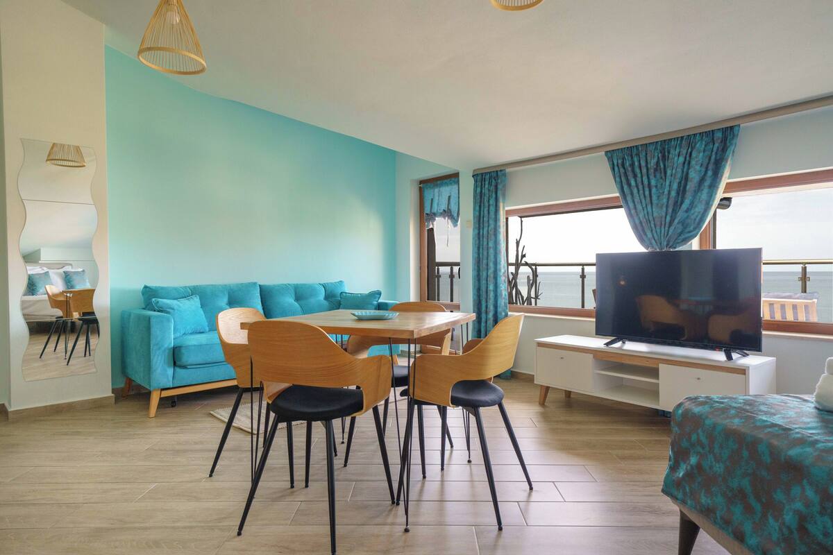 Un salon moderne avec un canapé de couleur turquoise, une table à manger en bois et des chaises, un téléviseur à écran plat et un balcon avec vue.