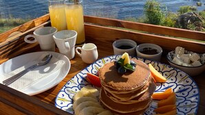 Bandeja de desayuno con panqueques, fruta y bebidas sobre una mesa de madera con vista al mar.