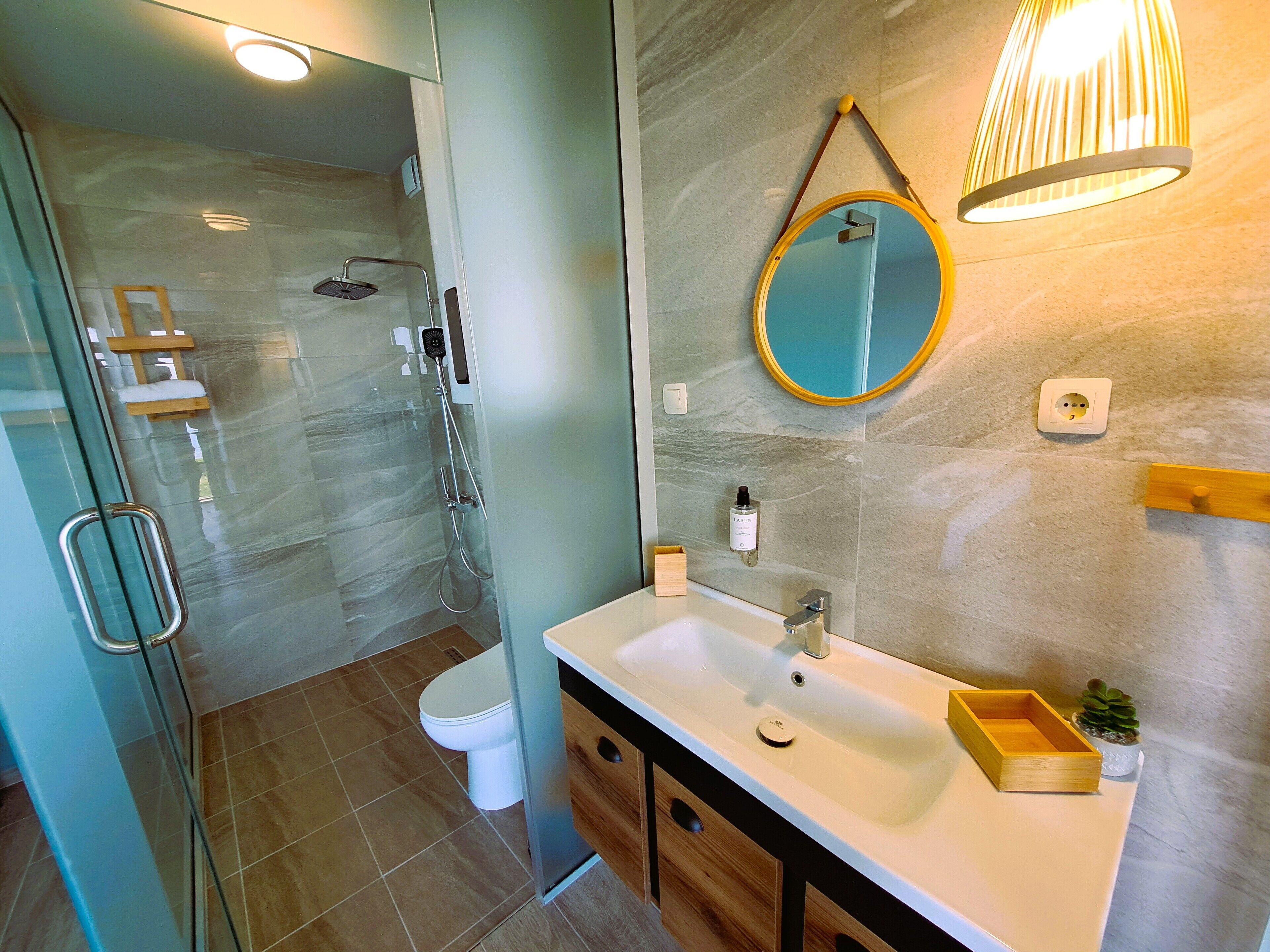 Een moderne badkamer met een glazen douchecabine, een wit wastafelmeubel met houten details, een ronde spiegel en een wandlamp.