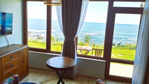 A room with a large window offering a view of the ocean, a wooden table, a TV, and a patterned sofa.