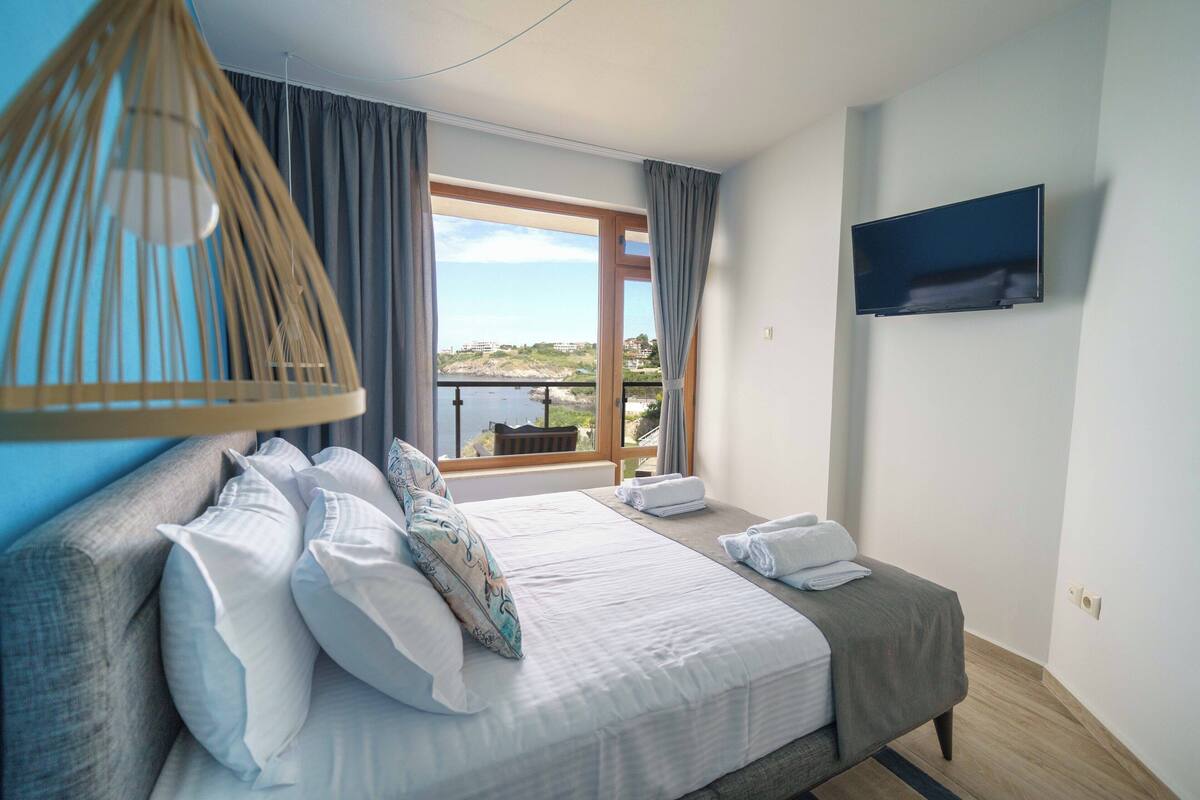Une chambre avec un grand lit, un téléviseur fixé au mur et une vue sur la mer par la fenêtre.
