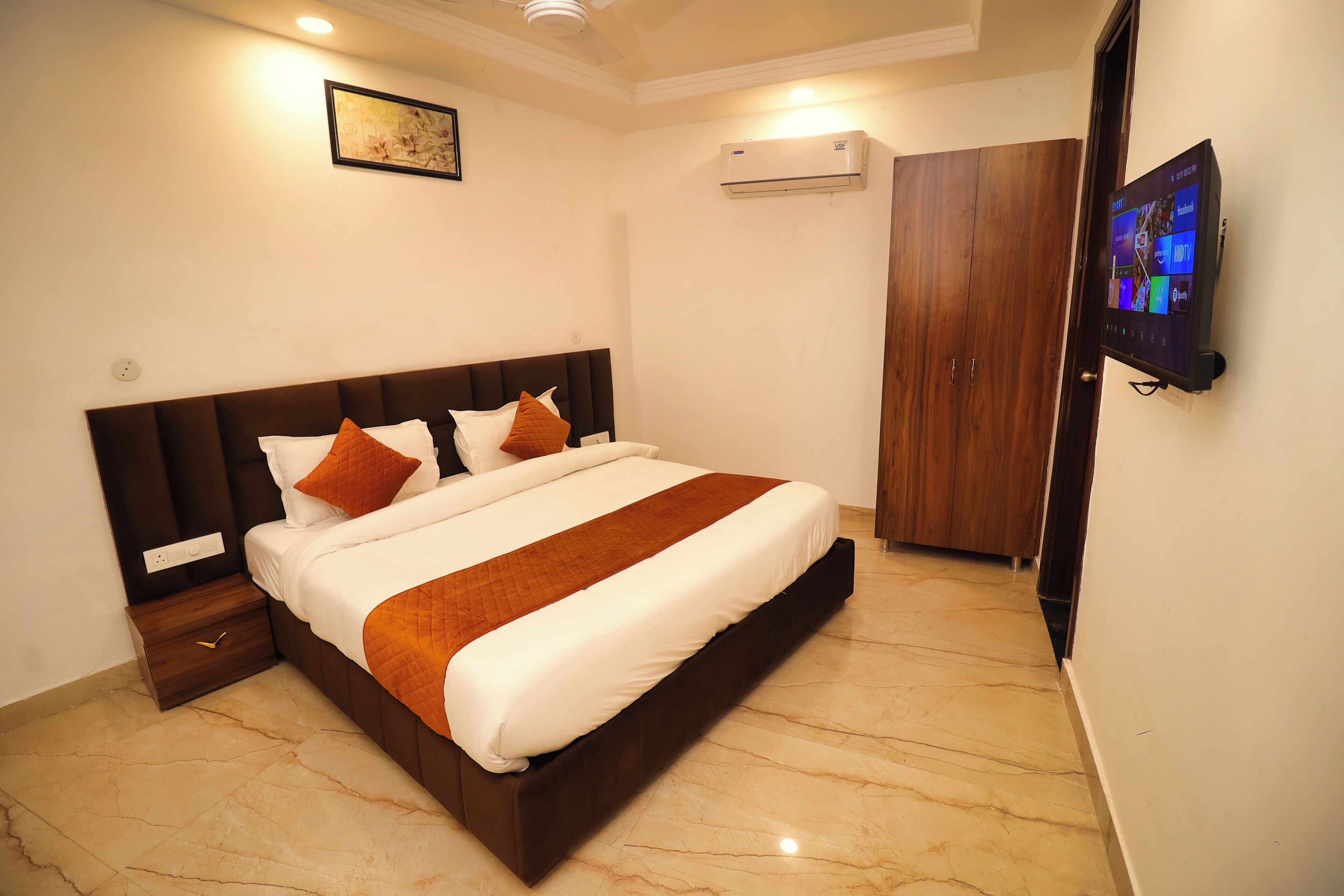 A neatly made bed with a brown headboard, a flat-screen TV mounted on the wall, and an air conditioning unit above it.
