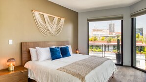 A modern bedroom with a large bed, a wooden nightstand, a wall-mounted macrame piece, and a balcony with a view of buildings and greenery. - Big PH Haven with rooftop, Condesa 2BR By Tasman (Ciudad de México)