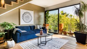 A modern living room with a blue sofa, a round coffee table, and a large window with a view of greenery. - Big PH Haven with rooftop, Condesa 2BR By Tasman (Ciudad de México)