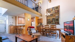 Un vaste espace de vie comprenant une table à manger, des chaises, un canapé et un téléviseur. Il y a également une cuisine avec des meubles en bois et un balcon doté d’une rambarde.